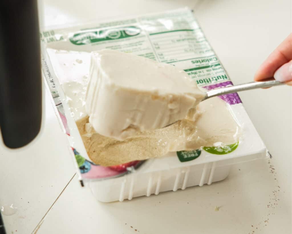 A hand scoops a large piece of soft tofu from an open plastic container with a spoon. The tofu looks smooth and creamy—perfect for making Vegan Crème Brûlée—and the nutritional label is partially visible on the packaging.